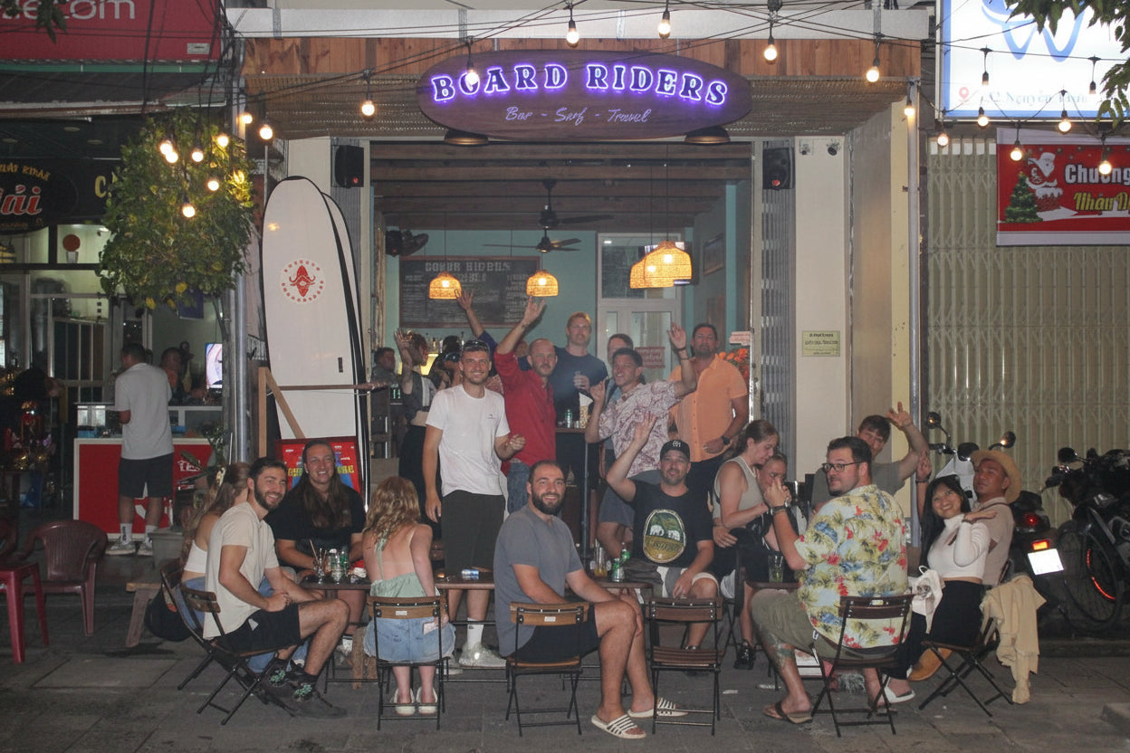  People sitting outside a bar named 'Board Riders' at night.